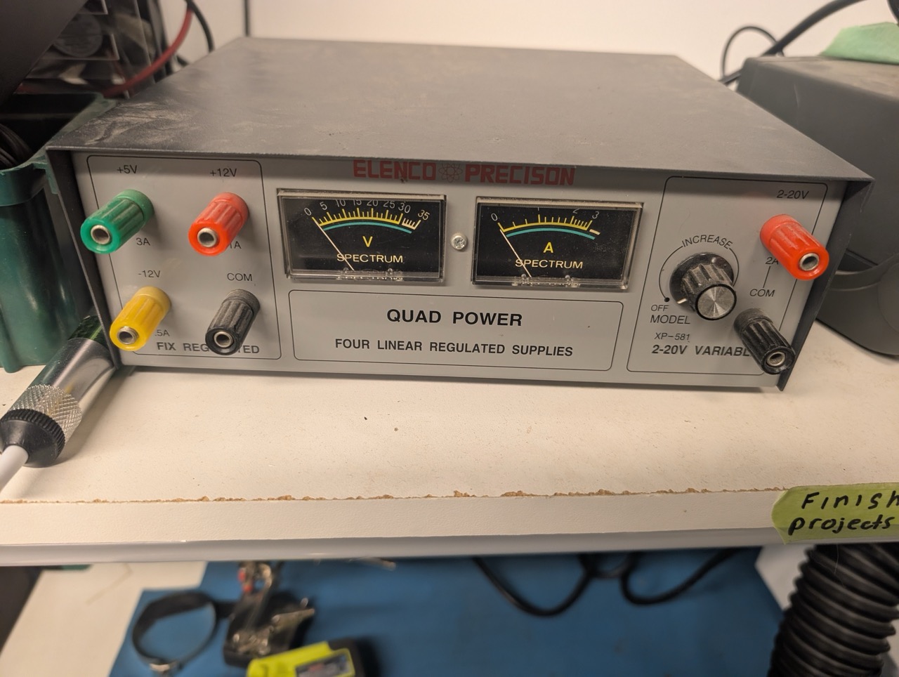 An older-looking horitzal-orientation power supply. There are 4 banana/binding posts on the left for +5V 3A (green), +12V 1A (red), -12V 0.5A (yellow) and COM (black). The center says "Elenco-Precision" and "Quad Power - Four Linear Regulated Supplies", with V and A analog meters. The right has a knob that says "Increase" above it, a red jack labelled 2-20V 2A, and a black COM terminal